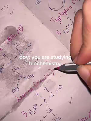 A close-up shot of a notebook page with handwritten biochemistry notes. A hand holding a pen is pointing at the notes, which include chemical structures and formulas. The notes are written with purple ink. The overall composition is focused and educational.