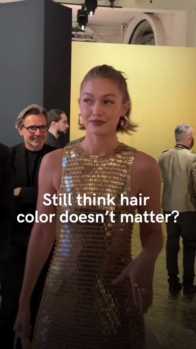 Gigi Hadid standing in front of a yellow wall, wearing a gold sequined dress. Other people are in the background.