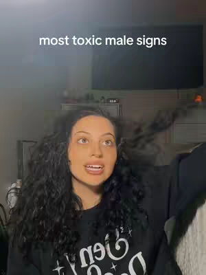 A young woman with long, dark curly hair and a black sweater with white graphic text looks upwards with an open mouth. The background is blurred, but includes a television and a hint of a bedroom setting. The image has a slightly desaturated, dim lighting.