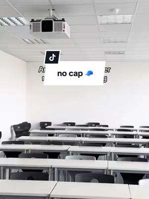 An empty classroom with rows of desks and chairs. In the foreground, a text overlay reading 'no cap' with a blue baseball cap icon. Above the desks is a ceiling with lights and a projector.