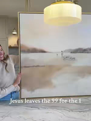 A woman in a light-colored sweater and jeans is holding up a large painting in a room. The painting depicts a serene landscape scene with a figure leading a group of sheep across a body of water.