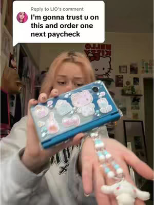 A woman with blonde hair holds up a blue DSi case decorated with stickers. In the background, there is a Hello Kitty poster and various items on a shelf, suggesting a bedroom or personal space. The image is brightly lit, with the woman’s face partially visible, focusing on the DSi case and her hands holding it. The person in the image has fair skin and is looking down slightly.