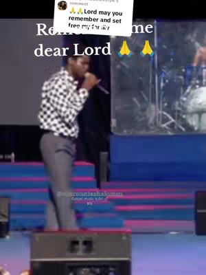 A man in a checkered shirt and gray pants stands on a stage, holding a microphone. The background includes stage lights. A text overlay reads, "Remember me dear Lord," with prayer hands emojis.