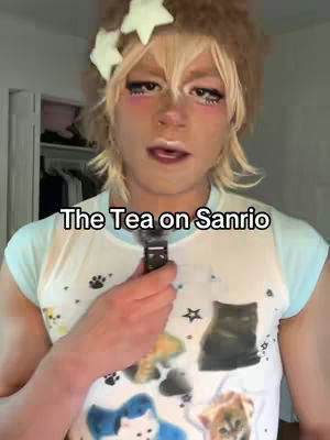 A person with light-colored skin, blonde hair and wearing makeup, is holding a small black object. The person is wearing a light blue t-shirt, with multiple colorful graphics including cats, and smiling. Text overlaid on the image says 'The Tea on Sanrio'. The background is a room with white walls and a partial view of a white closet.