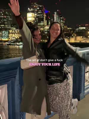 Two women are standing close together, smiling, with a backdrop of a city skyline. One woman has her arm raised. Text overlays read: 'The art of don't give a fuck ENJOY YOUR LIFE'. The color palette includes greens and blacks.
