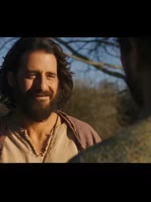 The image shows a man with a beard, wearing a tan and dark pink robe, smiling at someone off-screen. The background appears to be a natural setting with trees and soft lighting.