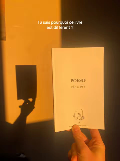 The cover image shows a hand holding a book with a white cover. The book's title is visible, and the upper right corner features text in French. The book is positioned against a wall with a shadow being cast on a plain surface from a light source. The hand casts a shadow on the wall.