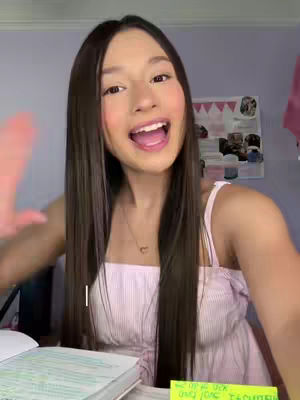 A young woman with long, dark hair is shown from the chest up, smiling at the camera with her mouth open, as if she is speaking. She has light pink lipstick on and is wearing a light pink dress. Her left arm is raised and her hand is slightly open. The background is a room with a purple wall, and various items are visible.
