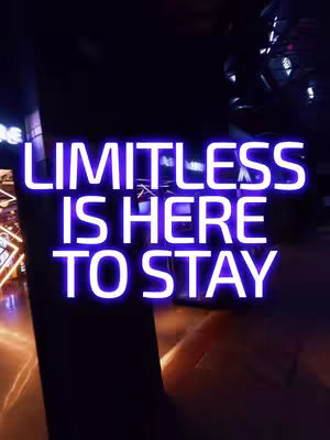 A dark, vertical image featuring bright, neon-style text that reads 'LIMITLESS IS HERE TO STAY'. The text is stacked in three lines, with a glowing purple and white color scheme. The background appears to be a dimly lit arcade or entertainment venue, with blurred lights and shapes suggesting activity. The lighting highlights the text prominently against the darker backdrop.