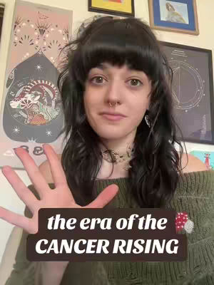 A woman with dark hair and a nose ring is centered in the frame, her right hand raised with fingers spread wide. She poses in front of a wall with framed art. A text overlay at the bottom reads "the era of the CANCER RISING".