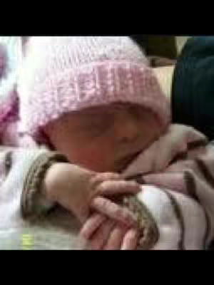 A close-up shot of a baby wearing a pink knitted hat. The baby's eyes are closed, and they are lying down with their hand up, and the camera angle is from the baby's perspective. The overall tones are soft and warm, with a focus on the baby's skin and the textures of the clothing.