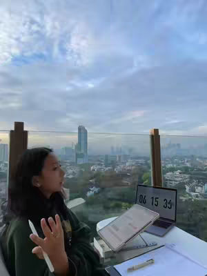 A young woman is sitting at a table on a rooftop with a cityscape background. She is looking up and to the left, appearing to be speaking or gesturing while holding a stylus in her right hand. On the table are a laptop displaying the time, a tablet, papers, and a pen. The sky is overcast, and the city below is in a slight haze.