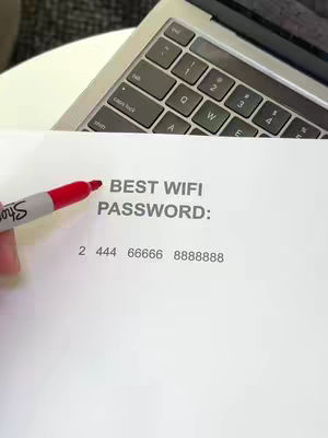 The image captured presents a casual indoor setting, with a laptop keyboard partially visible in the upper portion, indicating a workspace environment. Below, a sheet of white paper is centrally positioned, bearing the handwritten inscription 'BEST WIFI PASSWORD:' in black font. Below the text, potential numbers are listed as the password, with a Sharpie highlighter poised near the text, ready to underline it. The composition is simple, but the elements imply instructional context.