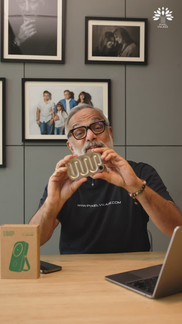 A man with glasses and a gray beard holds a rectangular metallic device with a wavy design in front of a gray wall. Behind him are framed photos on the wall. A laptop and a box labeled with "LOOP" are on a wooden table in the foreground.