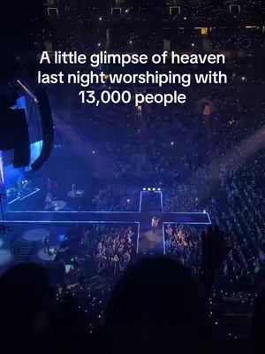 An overhead shot of a large concert venue, with a stage set up as a cross, bathed in blue and purple lights. The audience is densely packed with many people, creating a sea of lights.