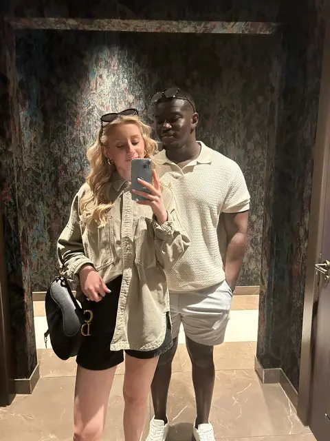 A couple takes a mirror selfie in a bathroom. The woman holding the phone is fair-skinned and has long blonde hair and sunglasses on top of her head. She wears a beige shirt and black shorts with a purse hanging from her shoulder. The man is black, with a beige polo and light-colored shorts. They are close together and smiling, and the background is a textured wall reflecting the overhead lighting.