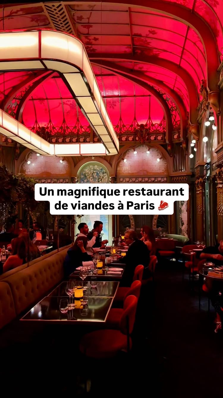 A shot inside a restaurant with red-tinted lighting. Tables are set with candles and glasses, occupied by individuals in conversation. The architecture has ornate details and arched ceilings, typical of a classic or upscale dining establishment.