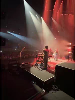 A dimly lit concert scene. A performer is on a riser stage, bathed in a spectrum of spotlights. The background reveals other performers and the audience, creating an immersive concert experience.