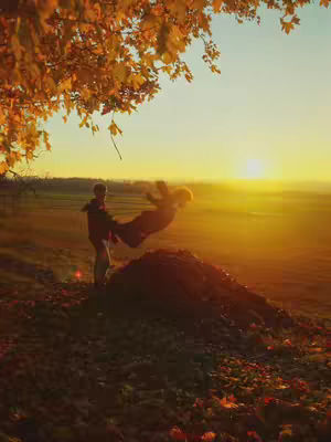 A man lifts a person in a pile of fall leaves with a golden sunset visible in the background.