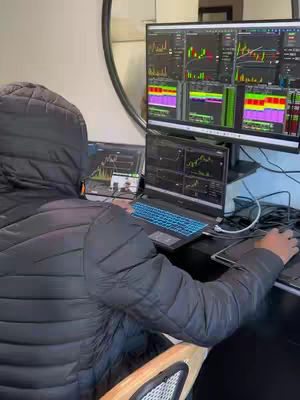 A person wearing a jacket sits at a desk with a large monitor displaying stock charts and a smaller laptop screen showing similar data. The person is working on a keyboard looking at the monitors.