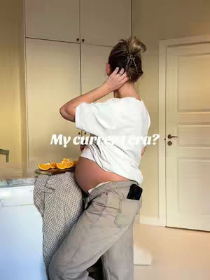 A woman stands mid-shot from a side angle, with her back to the camera. She is wearing a white t-shirt and beige pants, with one hand resting on her head. Her pregnant belly is visible. The background includes light-colored cabinets.