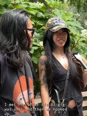 Two individuals standing side by side outdoors. On the left is a man with long dark hair, sunglasses, and a graphic t-shirt. On the right is a woman with long dark hair, a camouflage hat and a tattoo on her left arm; she is holding a microphone. Both are smiling. The background consists of green foliage.