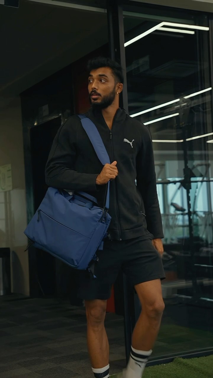 A man in athletic wear strides forward, carrying a blue messenger bag. The background suggests an office setting, with glass walls and artificial turf visible.