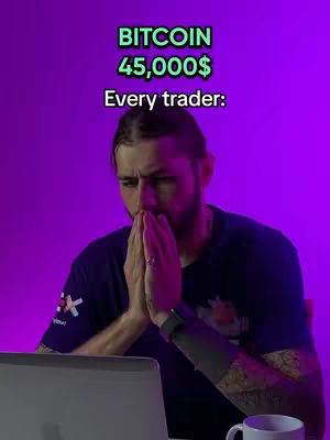 A man with a beard is captured in a medium shot, set against a vibrant purple backdrop. He wears a dark blue t-shirt with a logo on the left chest and a large tattoo partially visible on his left forearm. He is seated, with his hands clasped in prayer, a gesture that suggests contemplation or concern. A laptop rests in front of him, and to his right, a white mug is visible. Above the man, text overlays read 'BITCOIN' and '45,000$', followed by 'Every trader:' below, which is likely a hook.