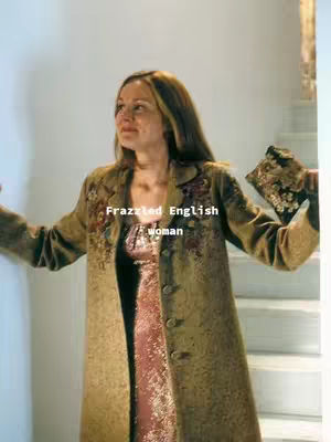 A woman stands indoors, facing slightly towards the left, with her arms outstretched. She wears a patterned coat over a pink dress. The background includes a staircase.