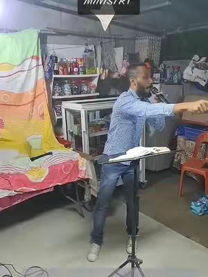 A man with a beard stands in a room speaking into a microphone. A sign that says MINISTRY is above and light shines from the top down, a book is on a music stand in front of him, and he points to the right.