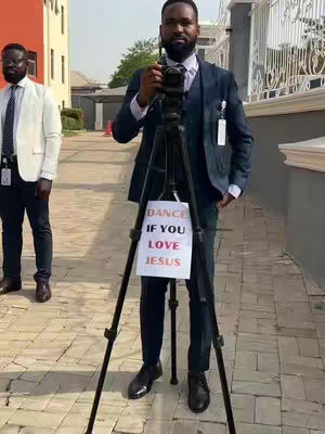 A man in a navy blue suit holds a camera on a tripod, with a sign on the tripod that reads "DANCE IF YOU LOVE JESUS." Another man is standing in the background wearing black framed glasses and a white suit.