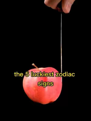 The image depicts a red apple suspended against a black background. A thin, light-colored string or thread extends from the top of the image, presumably holding up the apple. The apple is in focus and centrally positioned. Overlaying the apple is yellow text reading "the 5 luckiest zodiac signs".