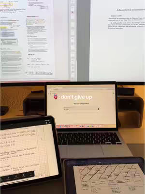 The image displays a laptop in the center, with a tablet on the left and another tablet on the bottom right, all open and showing content. A computer monitor rests on the desk above the laptop. The laptop's screen displays the text 'don't give up' in a creative font with a heart icon, suggesting a motivational theme. The lighting appears to be soft and diffused, creating an ambient and focused atmosphere.