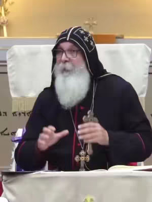 A man in religious attire. He is wearing a black hooded robe, glasses, and a white beard. A large cross hangs around his neck. The background is a blurred interior with a light-colored wall and other subtle details. The lighting is soft, creating highlights. The composition is centered on the man, with his hands gesturing.
