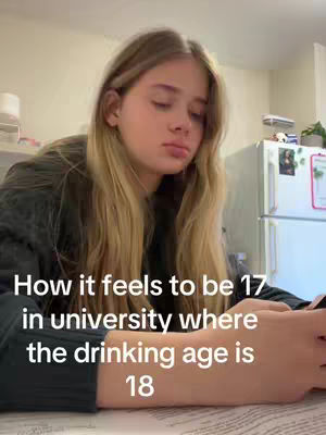 A young woman with long blonde hair is featured in a selfie-style shot. She appears to be indoors, possibly in a dorm room or a similar setting, as there's a white refrigerator and other furnishings in the background. The text overlay focuses on the challenges of being 17 and in university where the legal drinking age is 18.