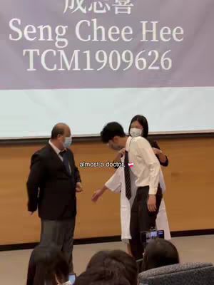 A video cover shows three people on a stage. In the foreground, two individuals are standing close, one in a white coat and the other in a dark suit and tie. A woman wearing a mask is placing the white coat on one of the men. The background features a banner with Chinese characters and English text.
