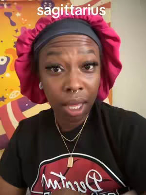 A close-up of a Black woman with a deep expression, wearing a burgundy head covering, a black shirt with a red and white design, and a gold necklace. Text in white arcs above the head is visible.