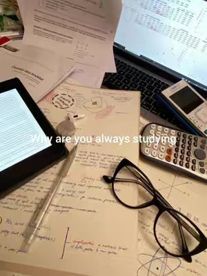 A desk with scattered papers, a laptop, a tablet, a calculator, and a pair of glasses. The overall tone suggests a study environment.