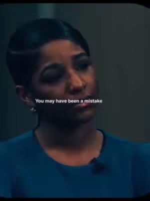A woman with short black hair, wearing a dark-blue top. She is looking slightly down and to the left, with a serious expression. The quote "You may have been a mistake" is overlaid on the image in white text. The background behind her is a plain wall, and the lighting is soft. The overall tone of image seems calm