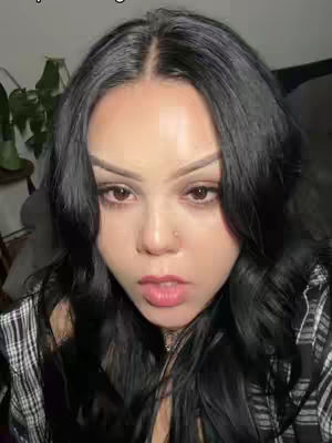 A close-up shot of a woman looking directly at the camera with a slight smirk. She has long, dark wavy hair and is wearing a black and white checkered shirt. The background is blurred, suggesting an indoor setting.