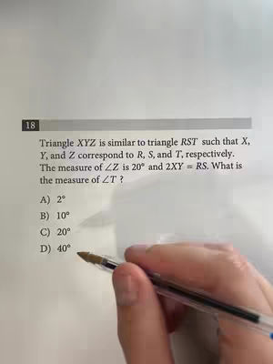 The image shows the top portion of a math problem printed on a sheet of paper. A hand holding a pen is visible in the foreground, pointing towards the problem. The text includes a math problem about angles and triangles. The background is a plain, light-colored surface.
