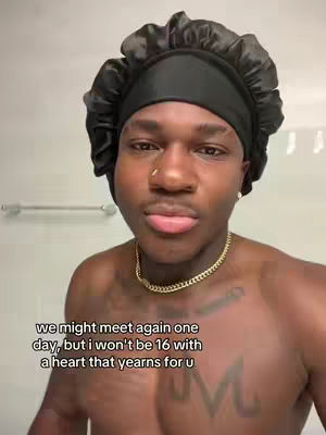 A selfie taken in a bathroom with a man with dark skin as the focal point. The man is wearing a black satin bonnet and a gold chain. Text overlayed on the image reads, 'we might meet again one day, but i won't be 16 with a heart that yearns for u.' In the background is a plain wall and a towel rack.