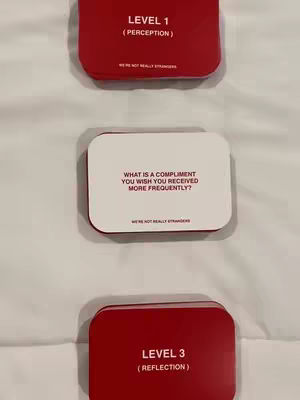 The image shows three cards from a card game, We're Not Really Strangers, arranged vertically on a white surface. The top and bottom cards are red with white text, while the middle card is white with black text. The top card reads 'LEVEL 1 (PERCEPTION)', and the bottom card reads 'LEVEL 3 (REFLECTION)'. The middle card poses the question 'WHAT IS A COMPLIMENT YOU WISH YOU RECEIVED MORE FREQUENTLY?'. The cards are slightly rounded at the corners.