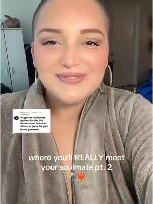 A woman with a shaved head is centered in the image, smiling at the camera. She wears hoop earrings and a gray robe. Text overlays read "where you'll REALLY meet your soulmate pt. 2". Below the text are ring and heart emojis.. A comment is overlayed on the left corner. The background is blurred and includes a cabinet.