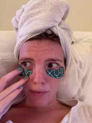 A close-up shot of a person in a white robe and a towel wrapped around their hair. They are wearing eye patches, looking in the mirror with a thoughtful expression. The person's finger is touching one of the eye patches.