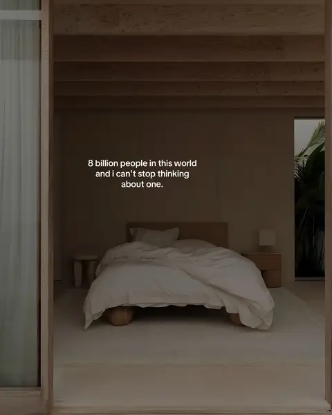 A serene interior shot framed through an open doorway. The central focus is a neatly made bed with white linens. The room is bathed in soft, diffused light, with a neutral color palette. The door frame and surrounding architectural elements are minimal in design, and a large window is visible. The text overlay reads: "8 billion people in this world and I can't stop thinking about one."