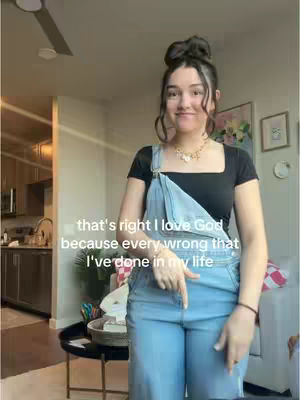 A young woman with dark hair styled in a bun stands in a well-lit living room with a kitchen visible in the background. She is wearing a black top, denim overalls, and a necklace. Text is overlaid on the image, and she is pointing downward.