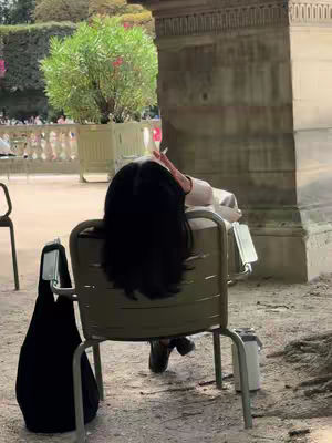 An outdoor shot shows a person sitting in a chair. The person is wearing a light-colored jacket and has long black hair covering their head, and the camera angle is positioned behind them. They are positioned amidst a scenic environment with foliage, hints of buildings, and other people.