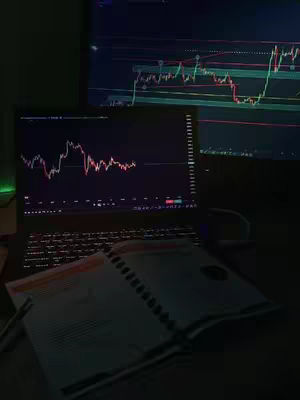 A dimly lit workspace with two computer screens displaying red and white stock charts. A laptop is open with a notebook and visible pen resting on the desk. The overall color scheme is dark, with screens as the primary light sources.