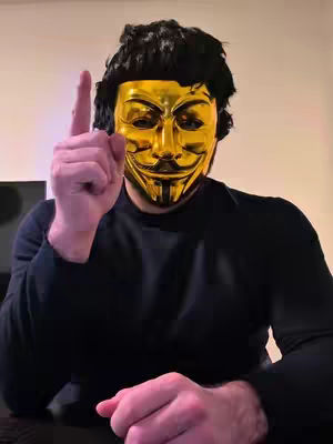 A person wearing a golden mask holds up an index finger, suggesting a point or emphasis. The person is wearing a black long-sleeved shirt, and only their hands and the top of their head are visible. The background has a neutral color. 
